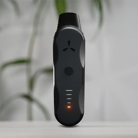 Vaporisateur à herbes AirVape XS Go debout sur une surface blanche avec LED orange allumée, plante floue en arrière-plan.